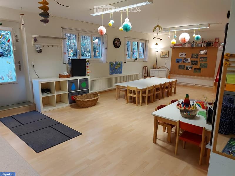 Brightly lit room with wooden flooring, large windows, and colorful decorations. Features a long table with chairs, a small shelf unit, a wicker basket, and various toys and educational materials.