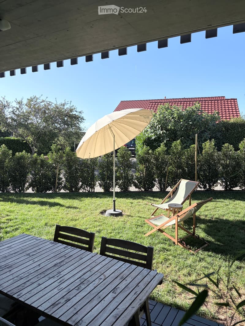 Garden area with a wooden table, chairs, and a large umbrella on a grassy lawn.