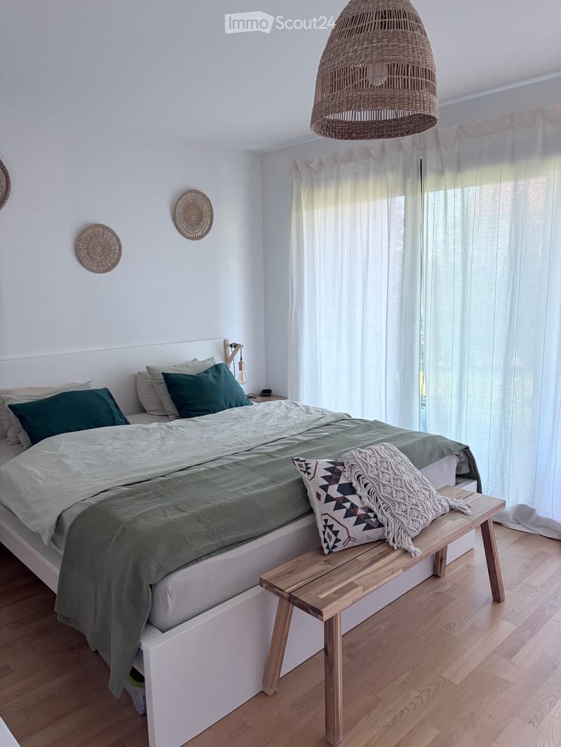 Modern bedroom with a white bed, green bedding, a wooden bench at the foot of the bed, and a woven pendant light hanging from the ceiling. The room features large windows with sheer curtains and a light wooden floor.