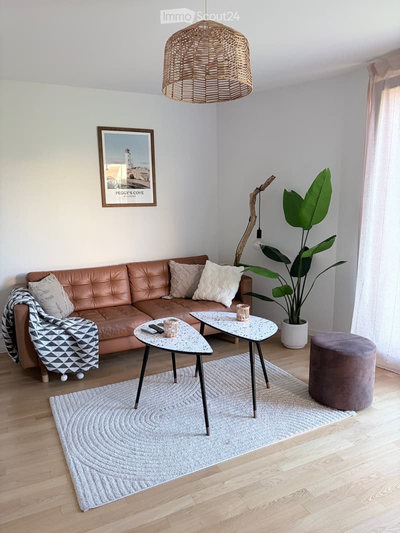 Modern living room with a brown leather sofa, a white coffee table, a grey rug, a large plant, and a hanging woven pendant light.