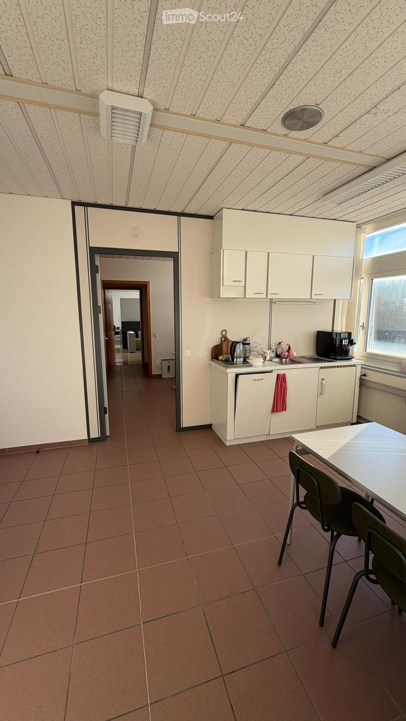 An empty room with brown tiles and white walls. It features a kitchen area with appliances like a coffee machine, kettle, and microwave. The room also has a table, chairs, and cabinets.