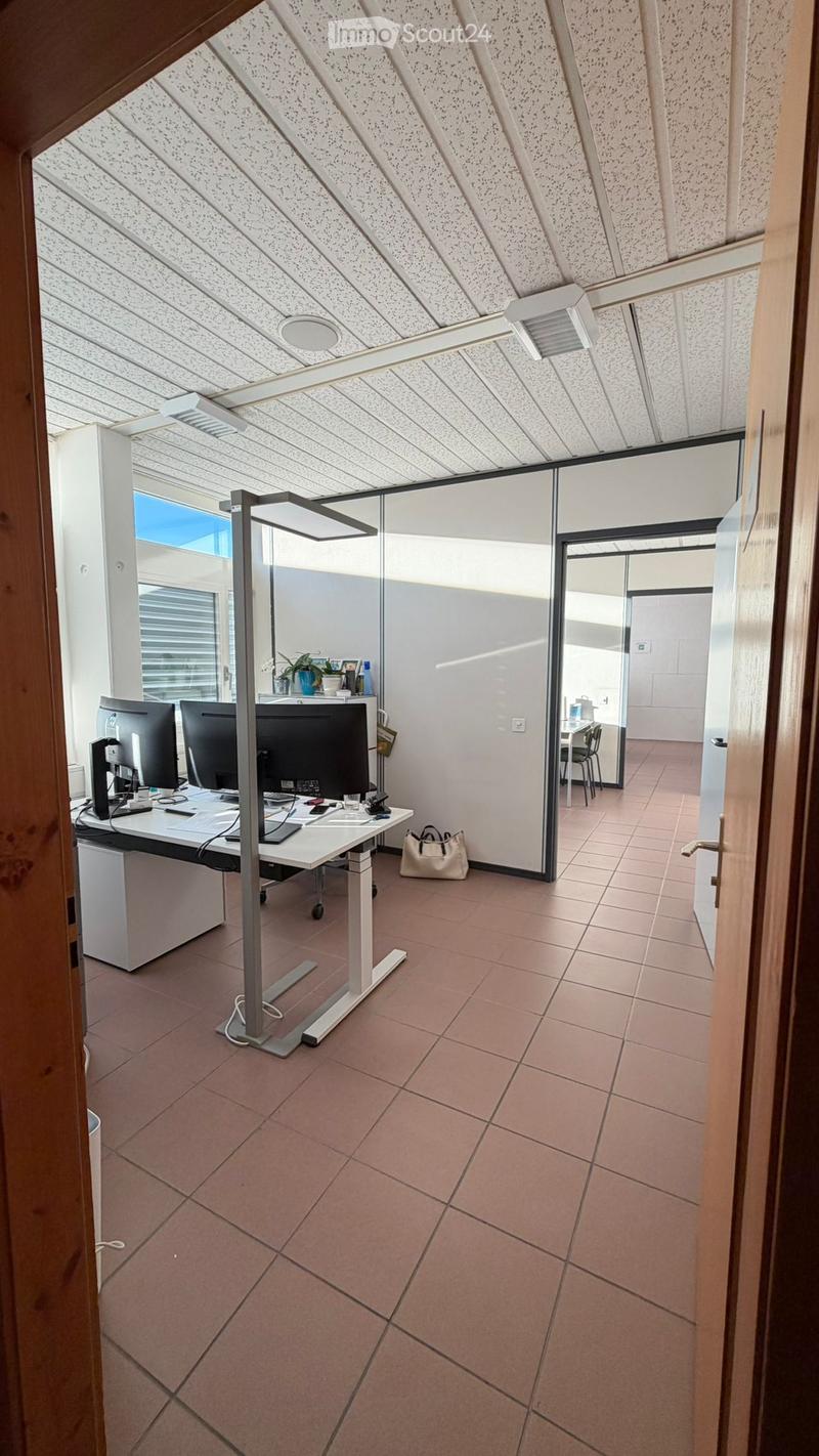 Spacious office space with a desk, monitors, adjustable height table, brown tiles on the floor, bright lighting, a bag, an office chair, and a glass door leading to a window.