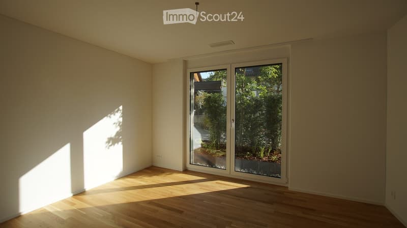 Empty room with light-colored walls and wooden flooring, featuring large glass sliding doors that open to a garden area.