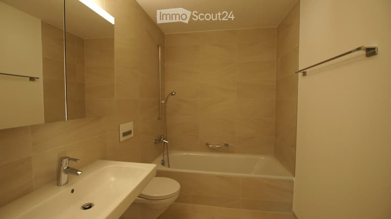 Modern bathroom with beige tiles, a white sink with a chrome faucet, a bathtub with a handheld showerhead, a toilet, and a large mirror with built-in lighting.