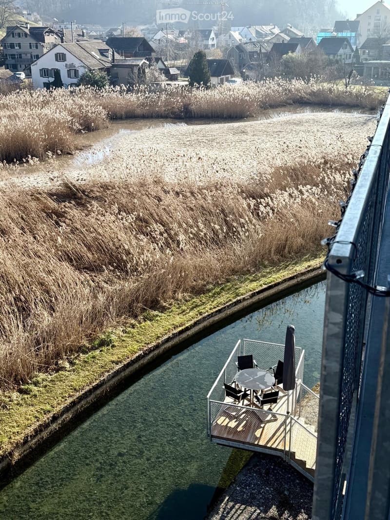 Balcony overlooking a river with a table, chairs, and an umbrella, surrounded by reeds and a small town in the background.