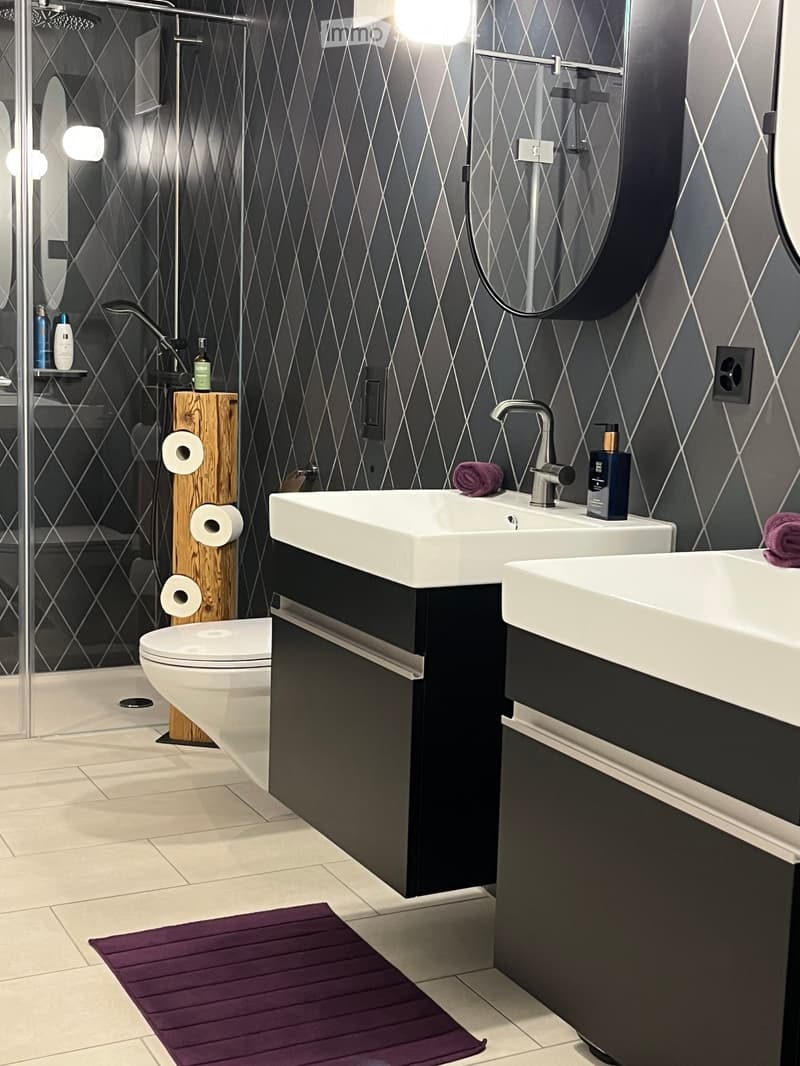 Modern bathroom with dark grey geometric patterned tiles, double sink vanity, toilet, shower area, and wooden toilet paper holder.