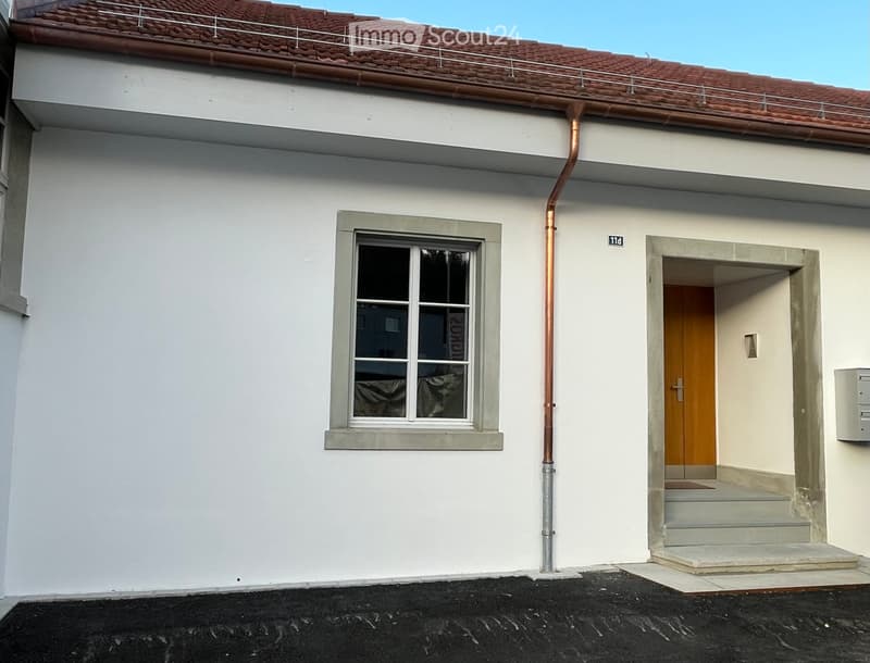 Two-story building with white exterior, red-tiled roof, and a small balcony on the roof. Features a window with a grey frame and a wooden door with a small step leading up to it.