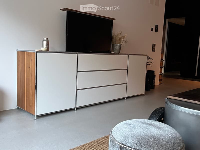 Modern living room with a grey ottoman, a sleek white and wood TV stand, a flat-screen TV, and decorative vases on the stand.