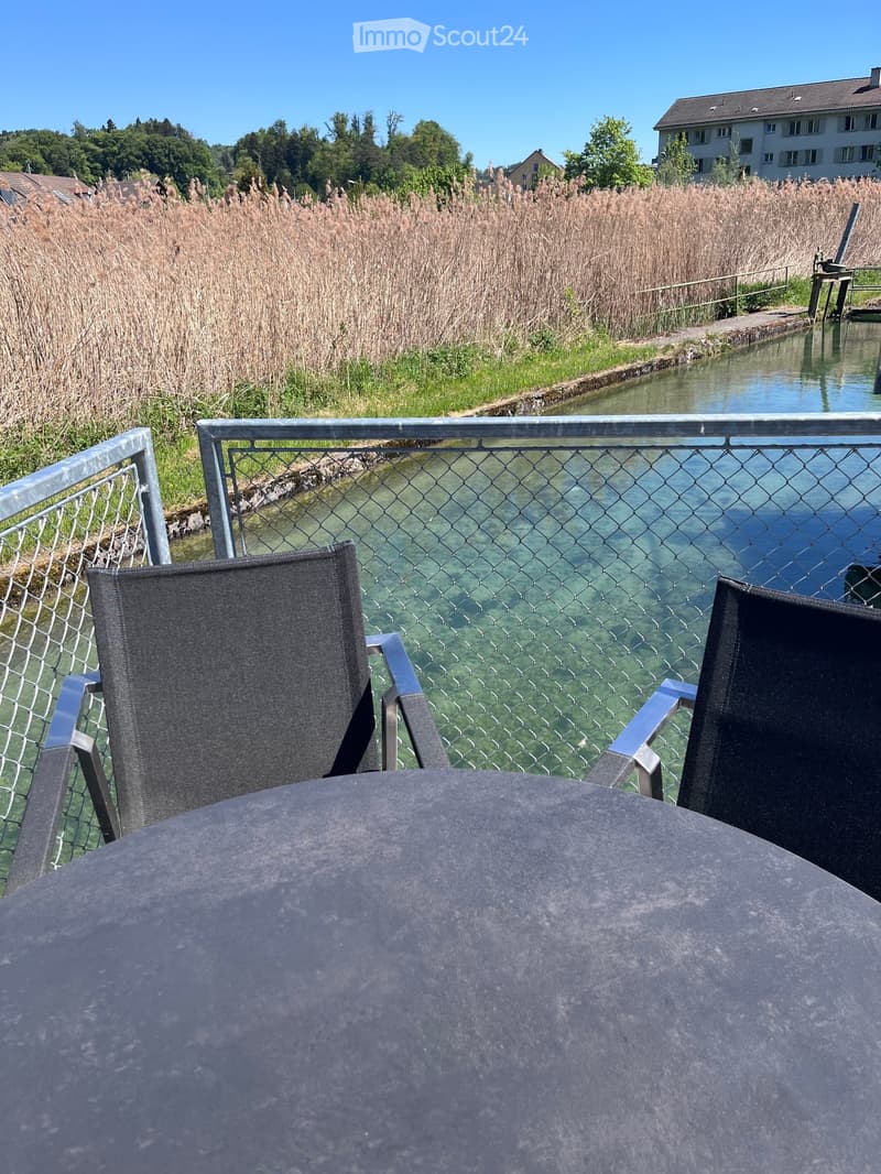 Balcony with two chairs and a table overlooking a canal with reeds and a distant house.