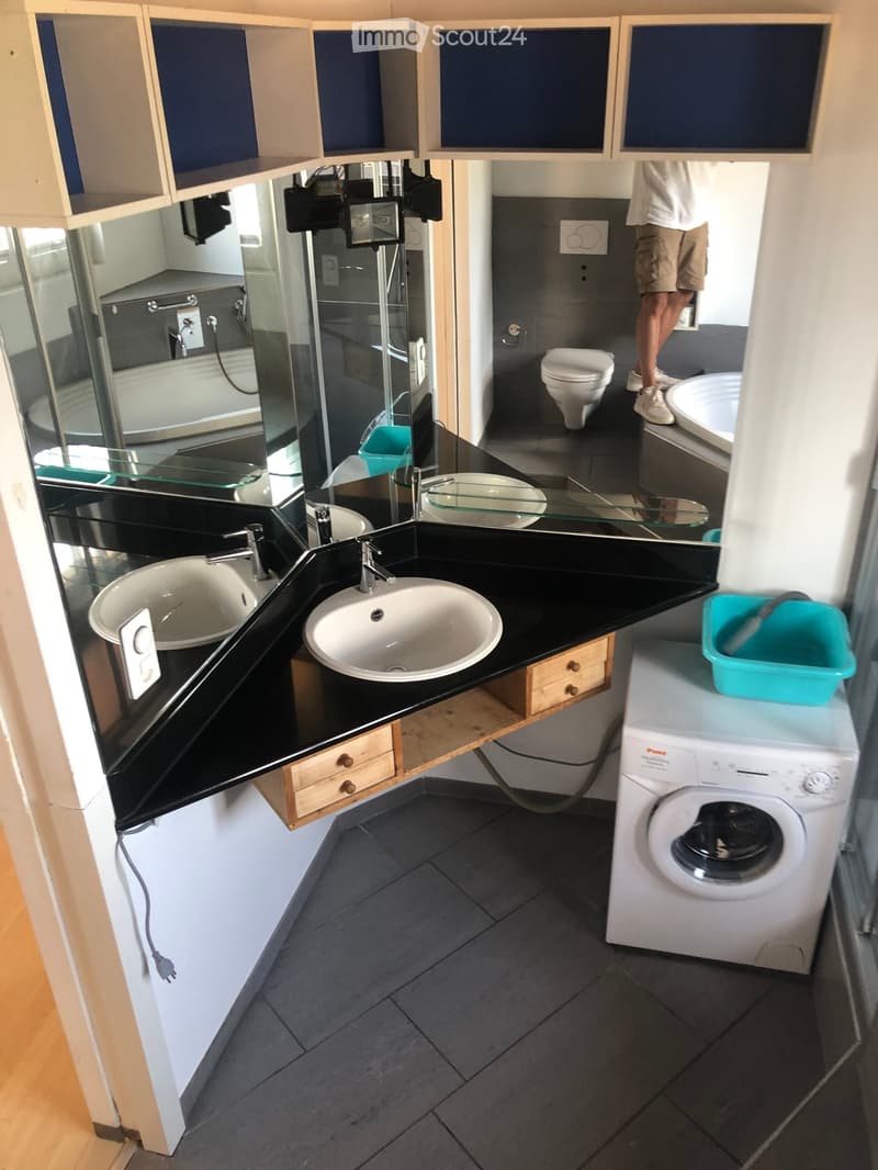 Modern bathroom with double sink, toilet, washing machine, and large mirror. Features dark countertops and tiled floor.