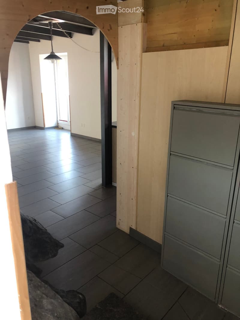 A partially furnished room with a refrigerator, tiled floor, and wooden walls. The space is open with visible beams and a hanging light fixture.