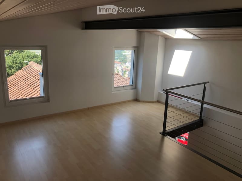 Empty room with wooden flooring, white walls, and large windows offering views of a tiled roof and surrounding area. A modern metal railing is visible, leading to an upper level.