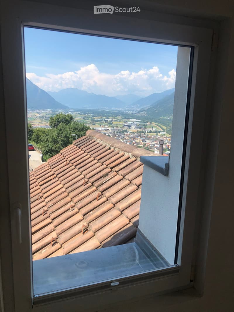 View from a balcony showing a terracotta-tiled roof, distant town, and mountain range under a clear blue sky.