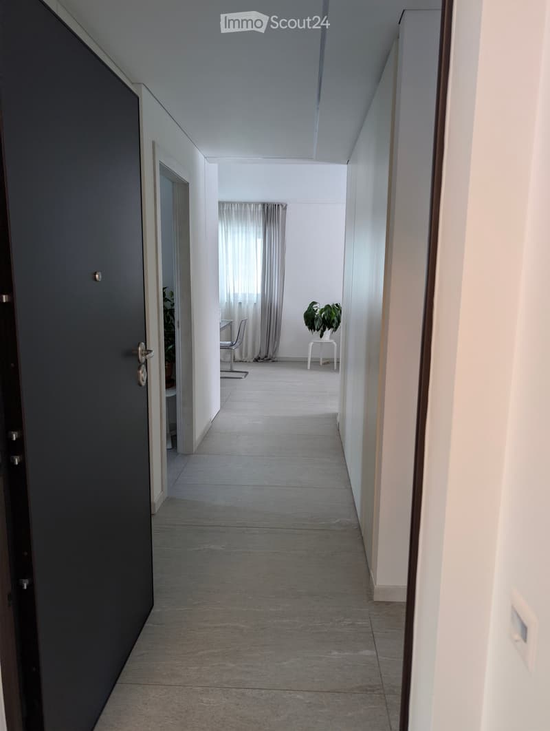 A long, narrow hallway with light-colored walls and a tiled floor. The hallway features a closed door on the left and an open doorway on the right leading to a room with a chair, small table, and plant.