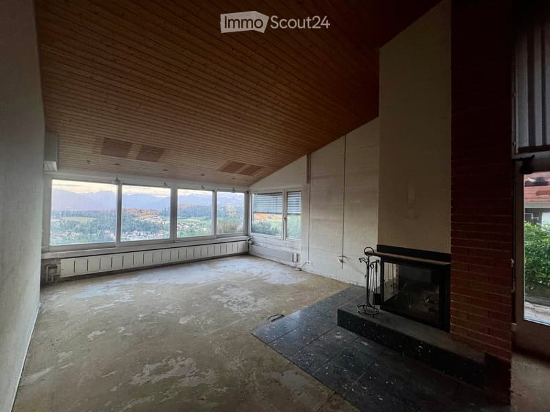 Empty room with wooden ceiling, large windows offering a scenic mountain view, and a brick fireplace with a small metal stand nearby.