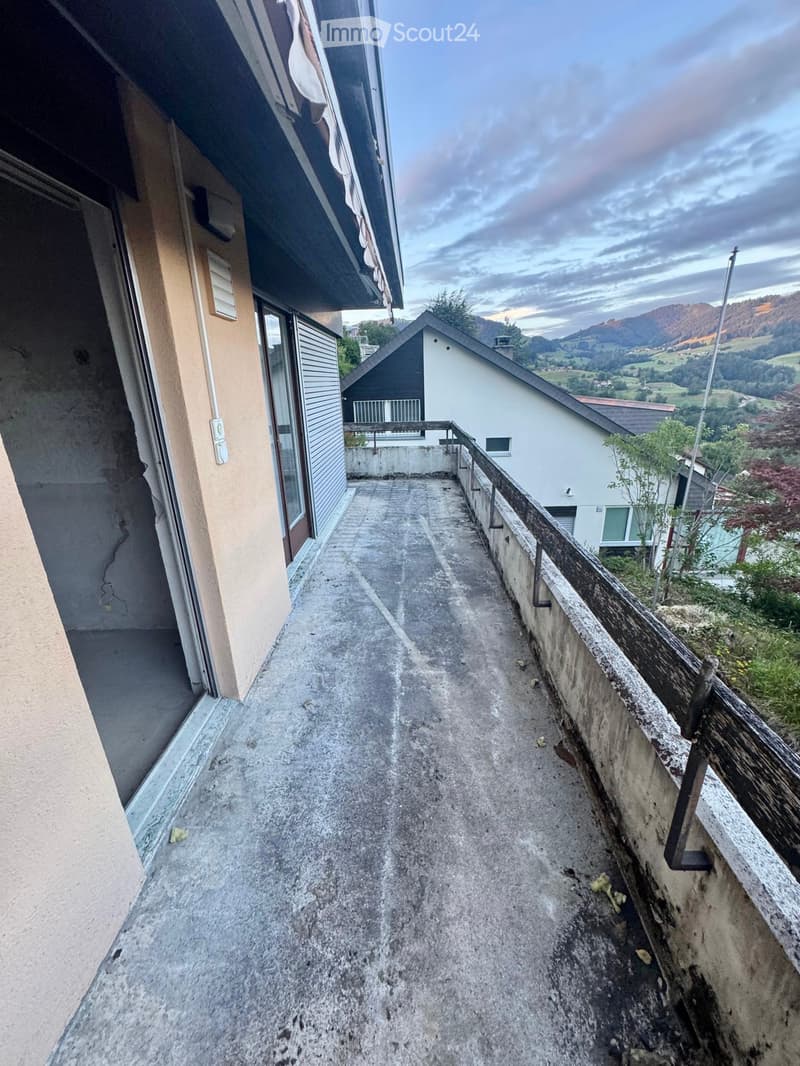 Concrete balcony with wooden railing, overlooking a rural landscape with houses and mountains.