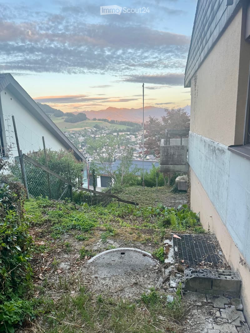 A sloped outdoor area with a concrete manhole cover, a metal grate, a small staircase, and a view of a town and mountains in the background.