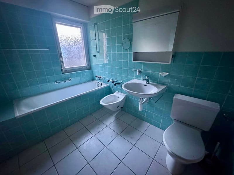 Bathroom with teal tiles, bathtub, toilet, sink, bidet, and a window with frosted glass.
