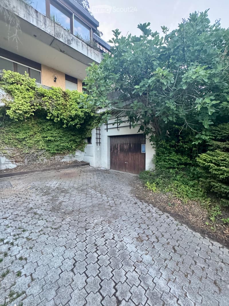 A multi-story building with a garage entrance, surrounded by greenery and a paved driveway.