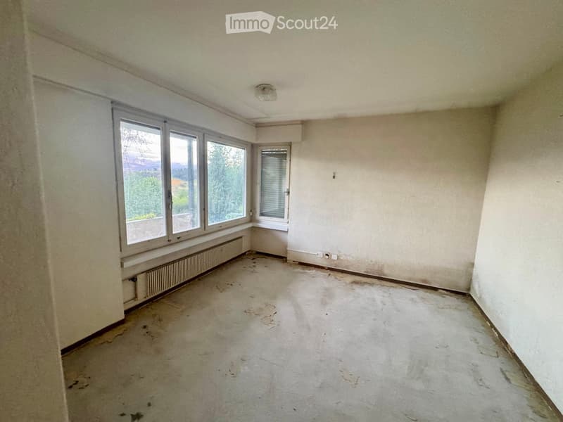 Empty room with bare walls, concrete floor, and large windows offering a view of greenery and mountains.