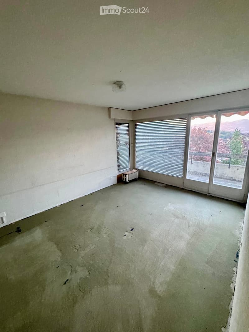 Empty room with unfinished concrete floor and walls, large windows with blinds, and a small radiator in the corner.