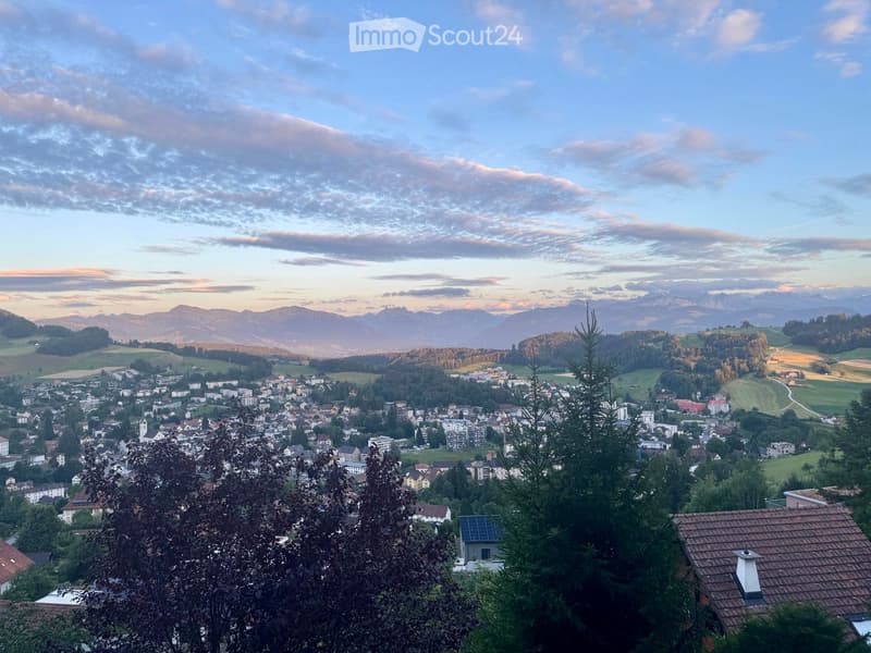 Panoramic view of a town nestled in a valley with surrounding mountains, featuring houses, trees, and fields under a partly cloudy sky.