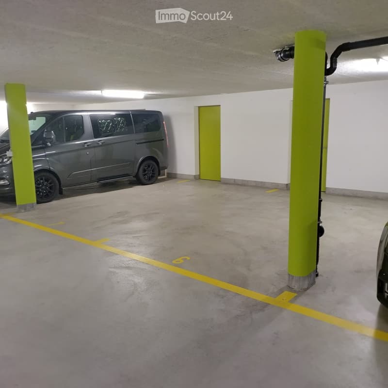 Underground parking area with a grey van parked, concrete floor, green pillars, and yellow parking lines.