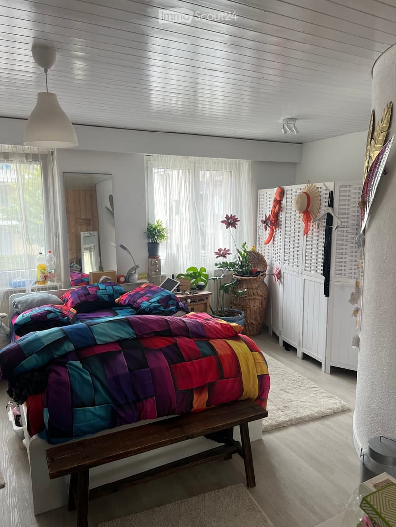 Bright bedroom with a colorful quilted bed, wooden bench at the foot, white walls, large windows with sheer curtains, and decorative plants. Includes a mirror, small table with items, and a hanging lamp.