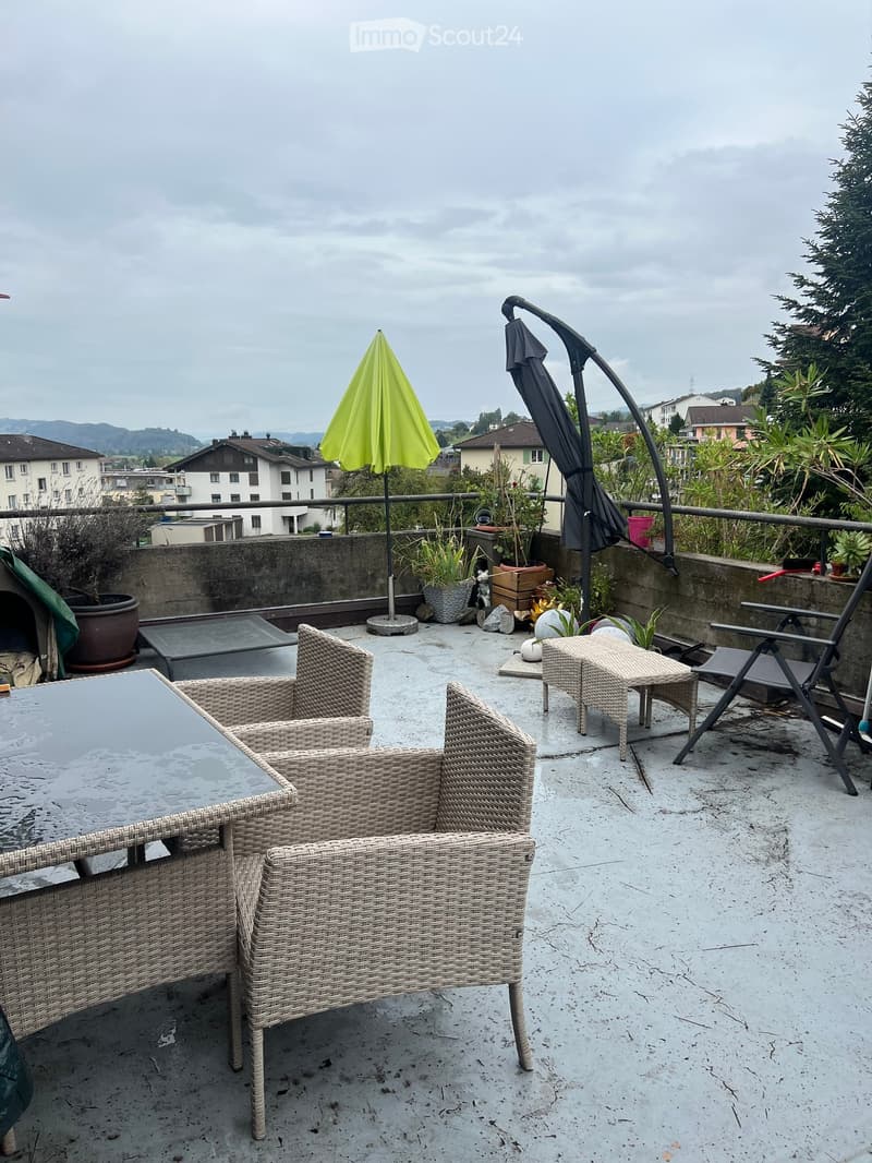 Balcony with wicker chairs, a glass table, and an umbrella, overlooking a town with houses and mountains.