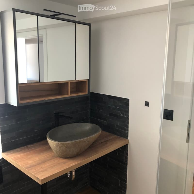 Modern bathroom with a wooden countertop, stone sink, black tiled walls, and a mirrored cabinet with wooden shelves.