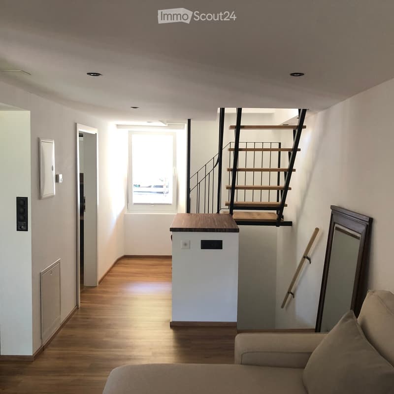 A modern, minimalist hallway with wooden flooring, a small table, a staircase leading upwards, and a sofa in the foreground.