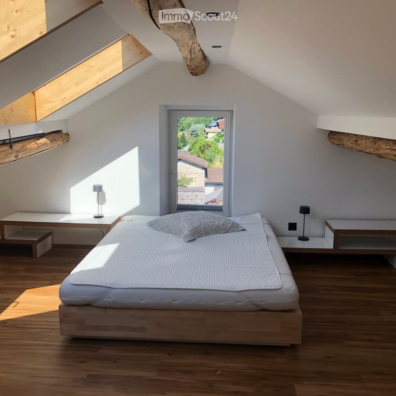 Bright, modern bedroom with wooden flooring, a large window offering a view, a neatly made bed with a decorative pillow, and two nightstands with lamps.