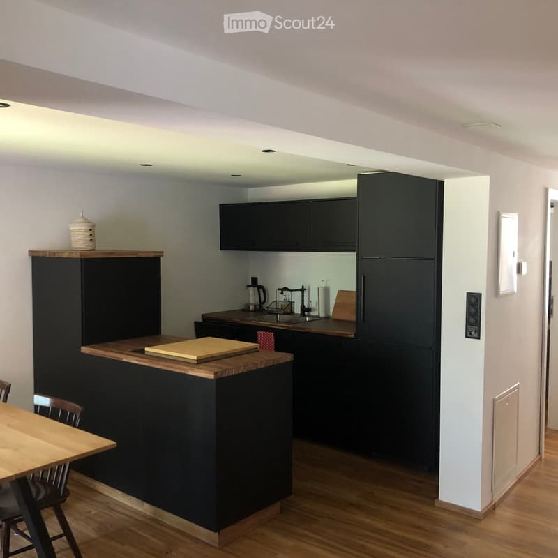 Modern kitchen with dark cabinets, wooden countertops, and a central island. Includes a refrigerator, sink, coffee maker, and a small dining area with a table and chairs.