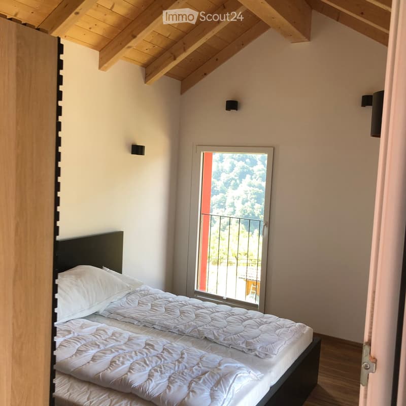 Cozy attic bedroom with a double bed, wooden slanted ceiling, and a large window opening to a balcony with outdoor view.