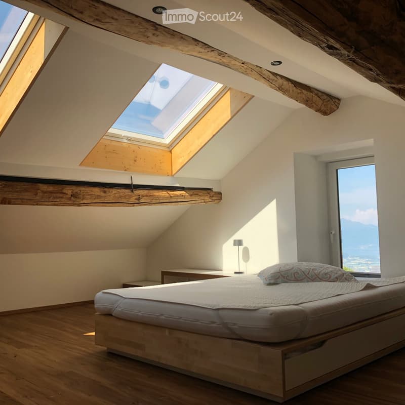 Bright, airy bedroom with wooden floors, exposed wooden beams, skylights, and a large window offering a scenic view.