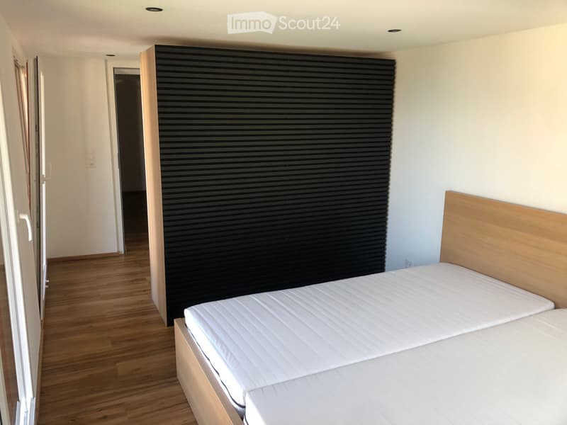 Minimalist bedroom with a single bed, white mattress, wooden headboard, and a black wall panel. Wooden flooring and white walls.