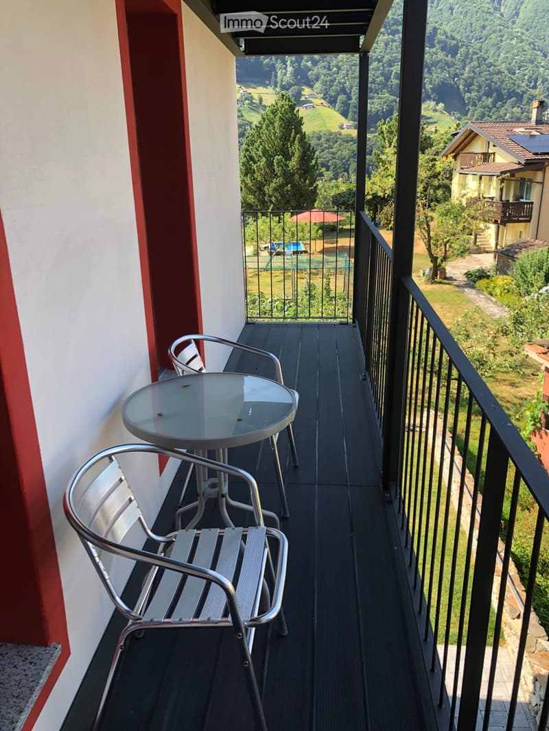 Balcony with a view of a green landscape, featuring a round glass table and two metal chairs.