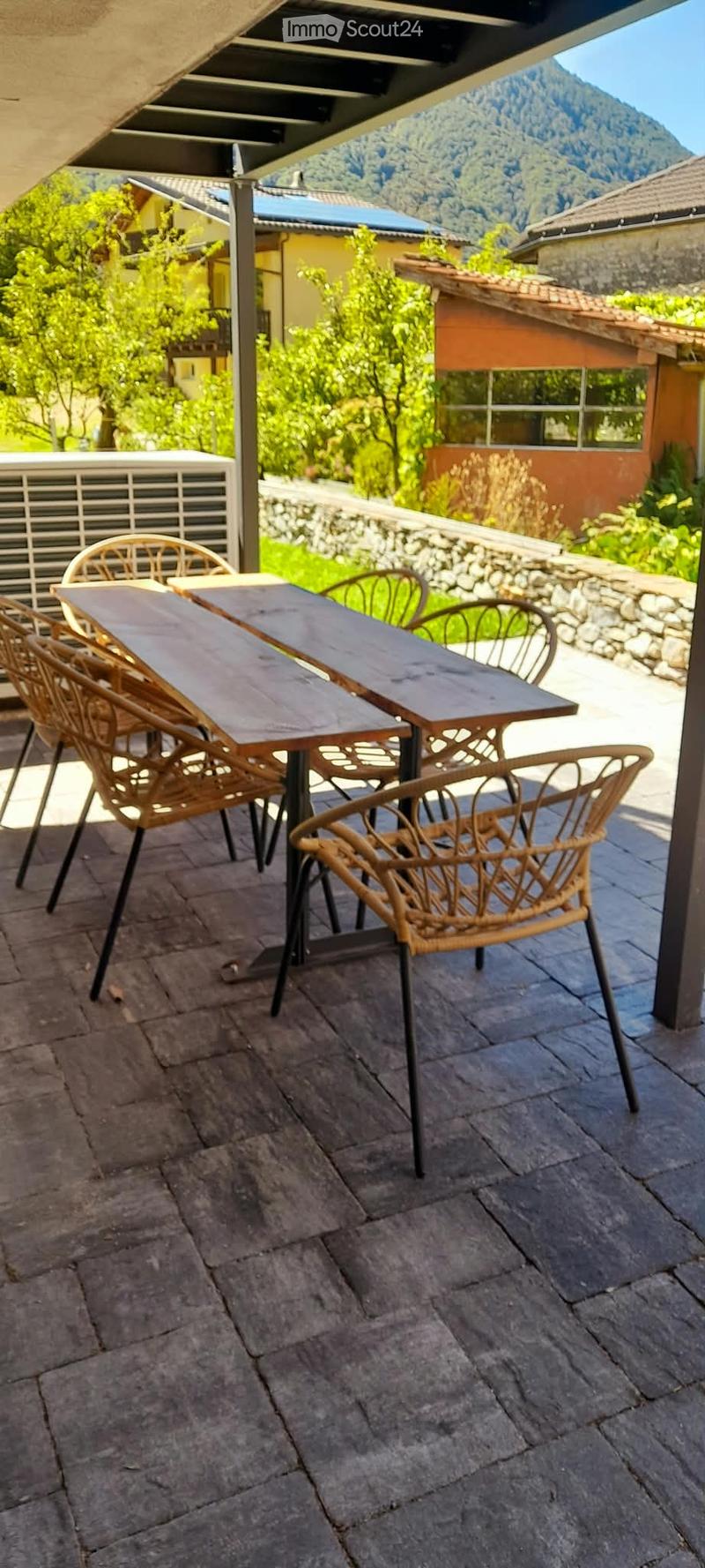 A stone-paved outdoor terrace with a wooden table and several wicker chairs, overlooking a scenic mountainous landscape with neighboring houses and greenery.