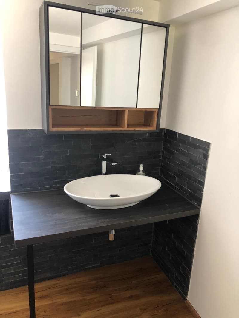 Modern bathroom with a white sink, dark wooden countertop, black tiled walls, and a large mirror with wooden shelves.