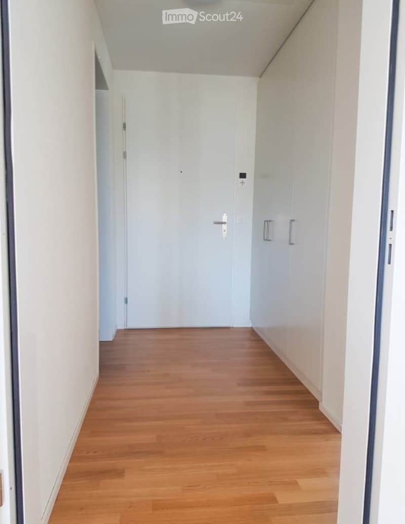A clean, modern hallway with light wooden flooring and white walls. Features include a closed door on the right and an open doorway on the left, with a small control panel near the ceiling.