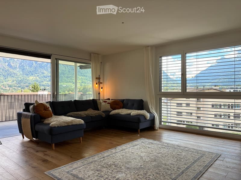 Bright living room with wooden flooring, large windows offering mountain views, a dark blue sectional sofa with throw pillows and a blanket, and an area rug.
