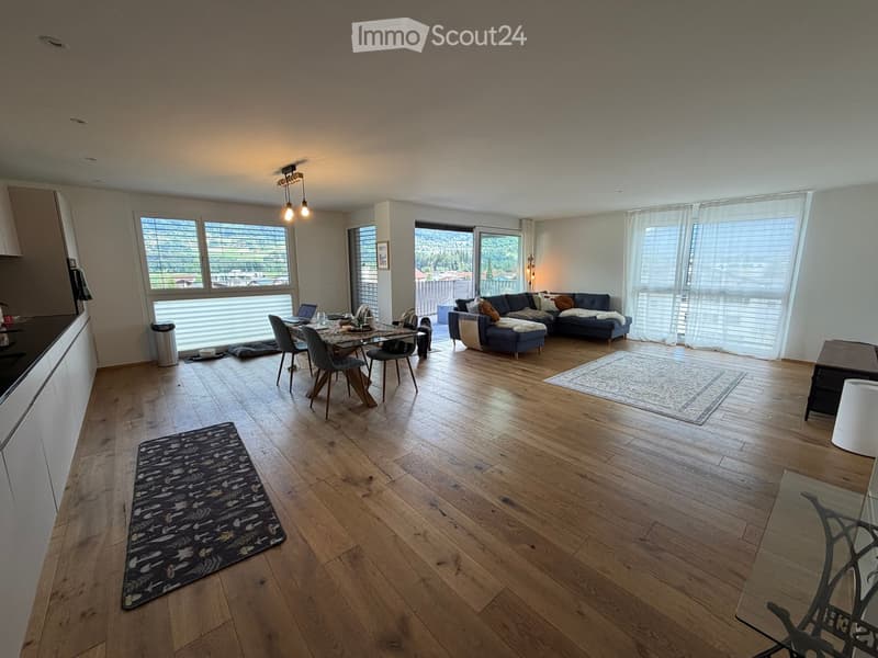 Open-plan living and dining area with wooden flooring, large windows, a sofa, dining table, and chairs.
