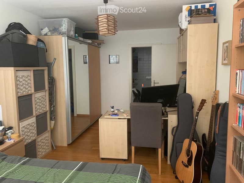 A furnished room with a bed, desk, chair, guitar, and storage cabinets. The room features wooden flooring and a hanging light fixture.