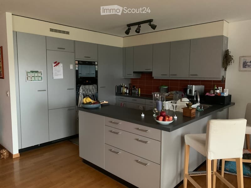Modern kitchen with grey cabinets, black countertop, and various appliances including a refrigerator, oven, and coffee maker. A stool is placed near the island, and a bowl of fruit is on the counter.