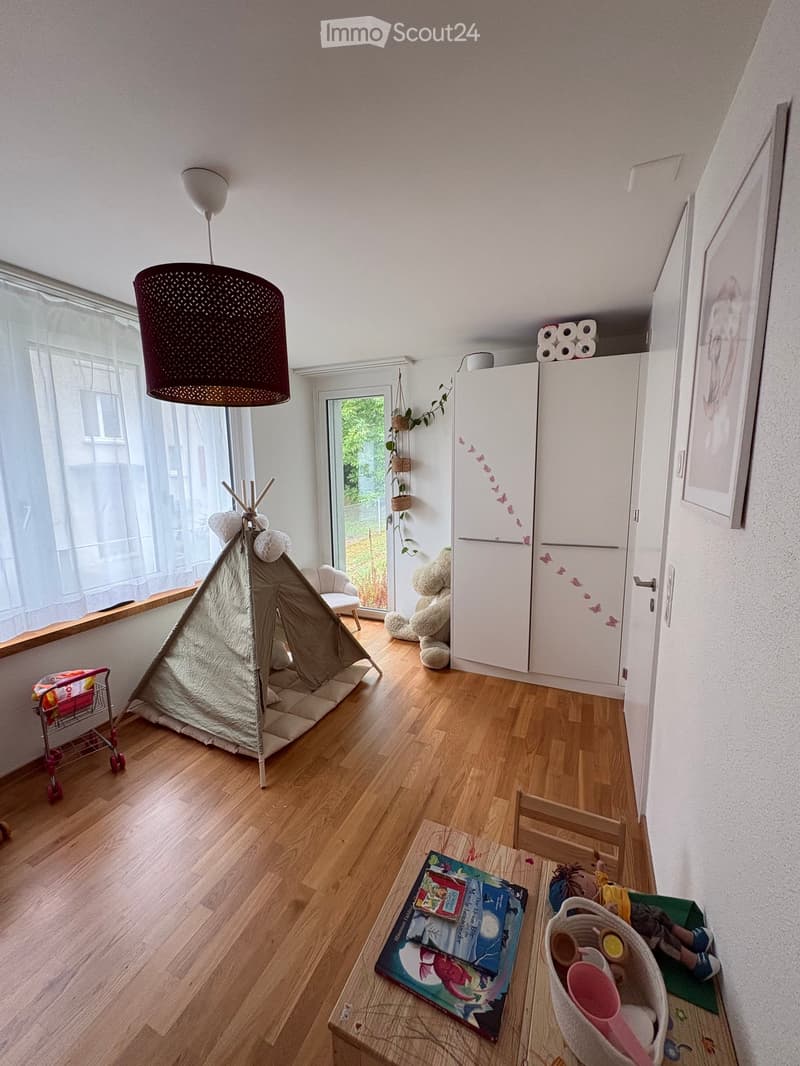 A well-lit room with wooden flooring, featuring a small table with books and cups, a toy tent, a teddy bear, a refrigerator, and a hanging plant near the window.