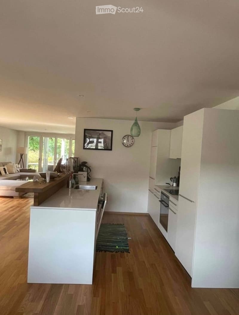 Modern kitchen with white cabinets, wooden flooring, and a central island. Features a sink, oven, refrigerator, and a small seating area with a sofa and coffee table.