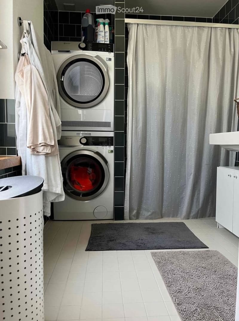 Modern bathroom with a stacked washer and dryer unit, a shower curtain, towels, cleaning supplies, a trash bin, and a bathroom cabinet.