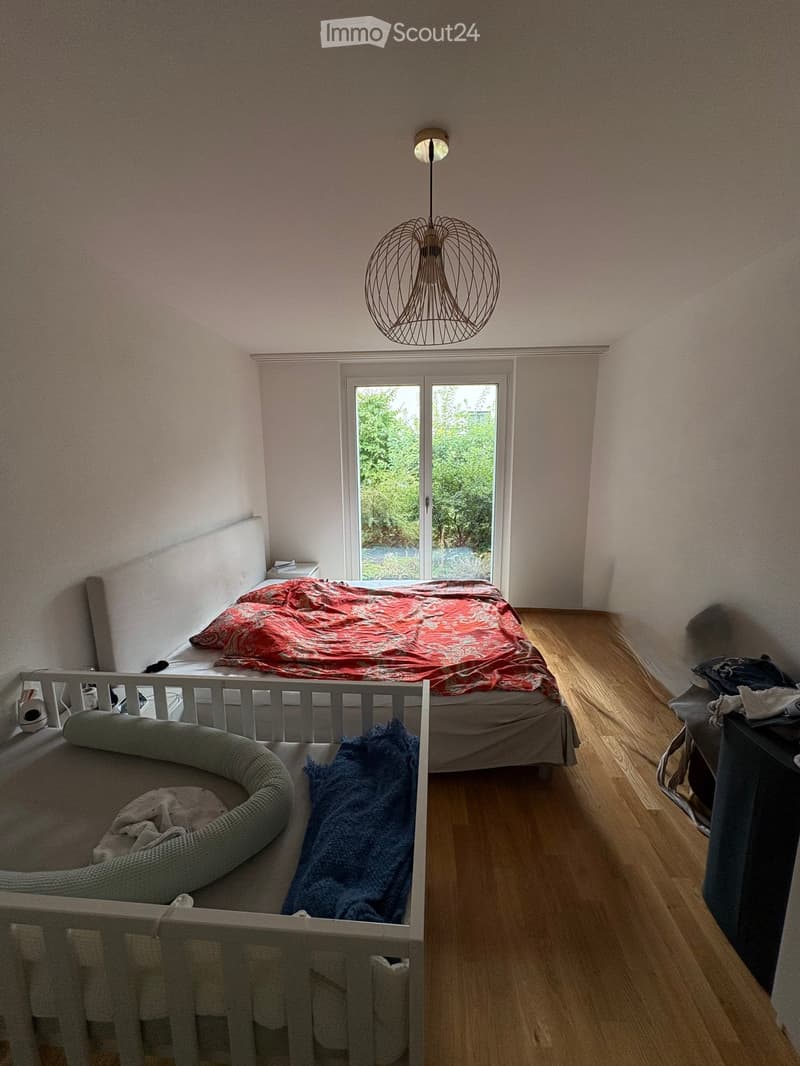 Cozy bedroom with a double bed, a cat bed, a nightstand, and a modern hanging lamp. The room features wooden flooring and large windows with a view of greenery.