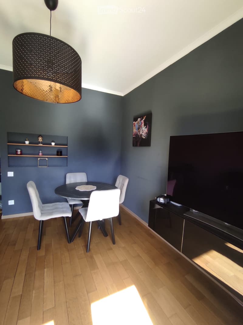 Modern living room with dark grey walls, wooden floor, round black dining table with four white chairs, large flat-screen TV on a stand, decorative wall shelves, and a large pendant light fixture.
