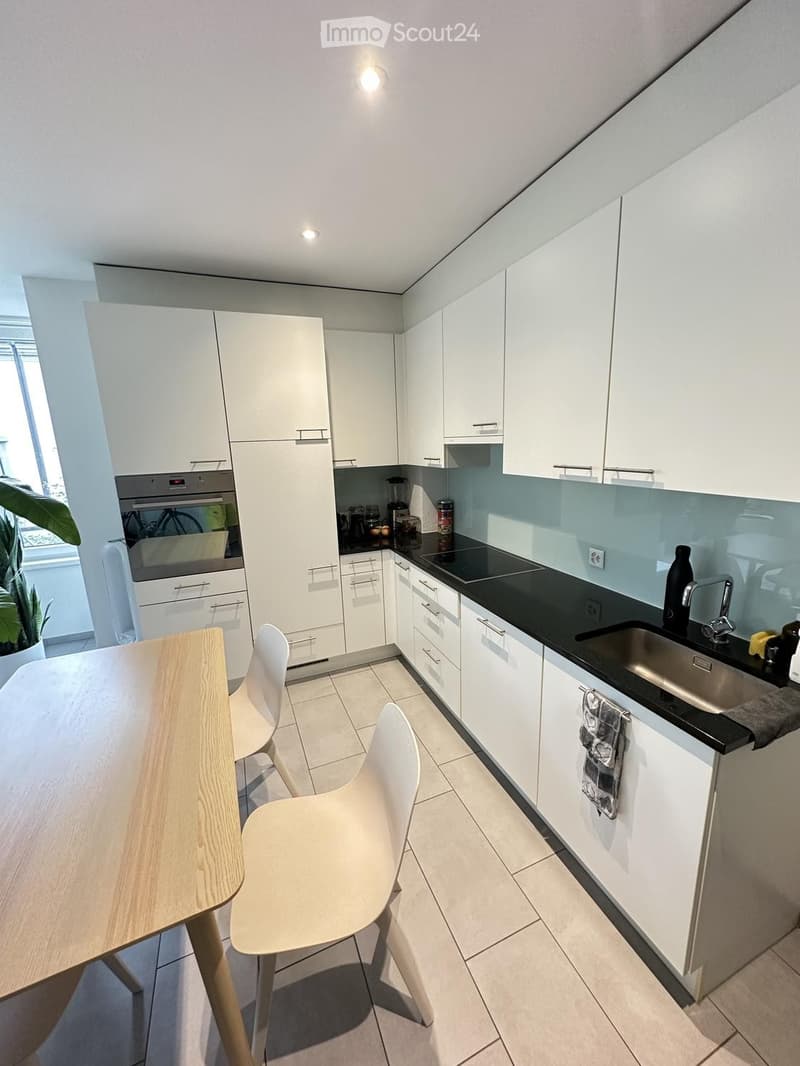 Modern kitchen with white cabinets, black countertops, a wooden dining table with white chairs, and various appliances including an oven, sink, and blender.